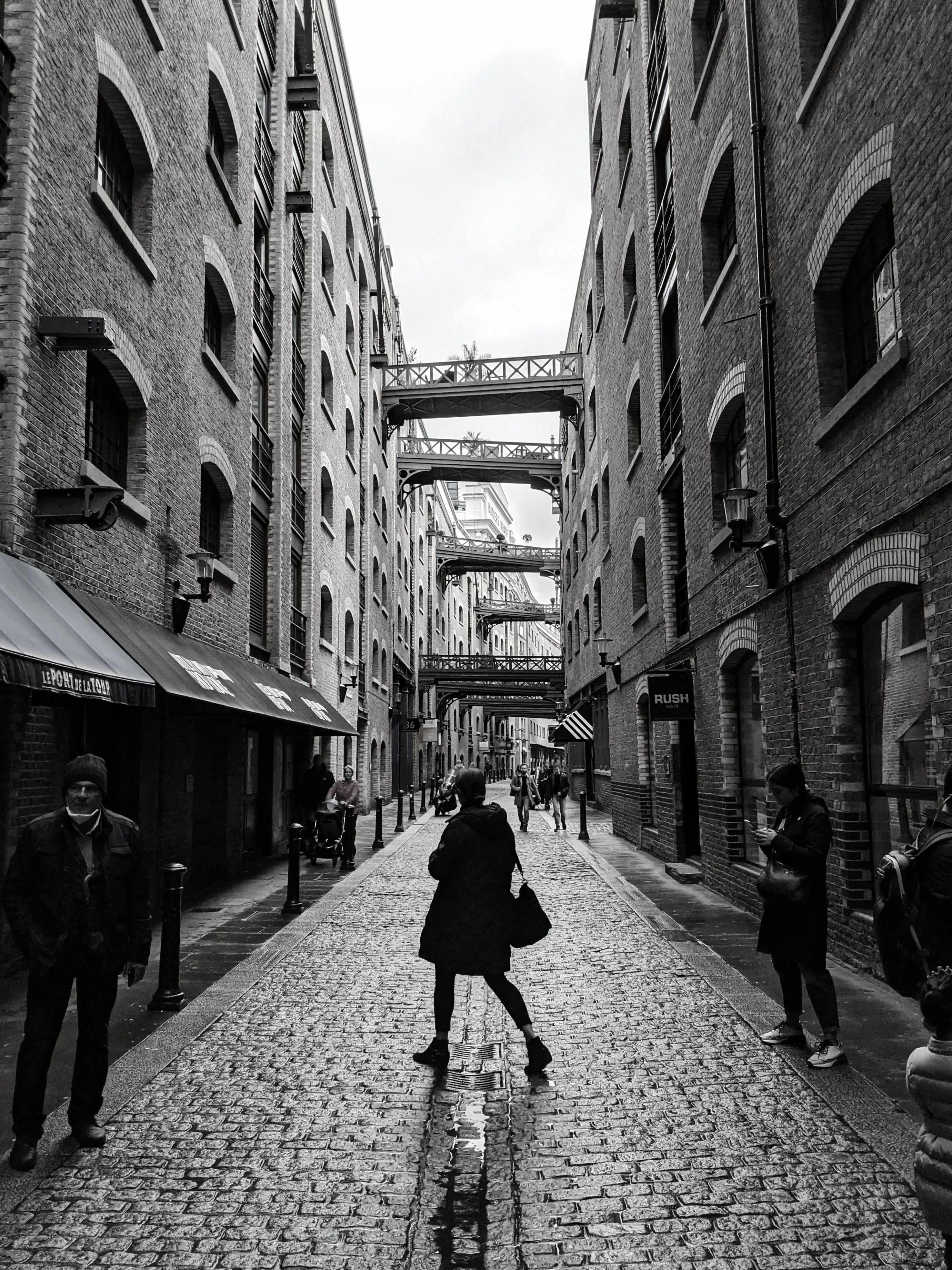 Black and white photo of a figure walking down a wet cobbled street between warehouses
