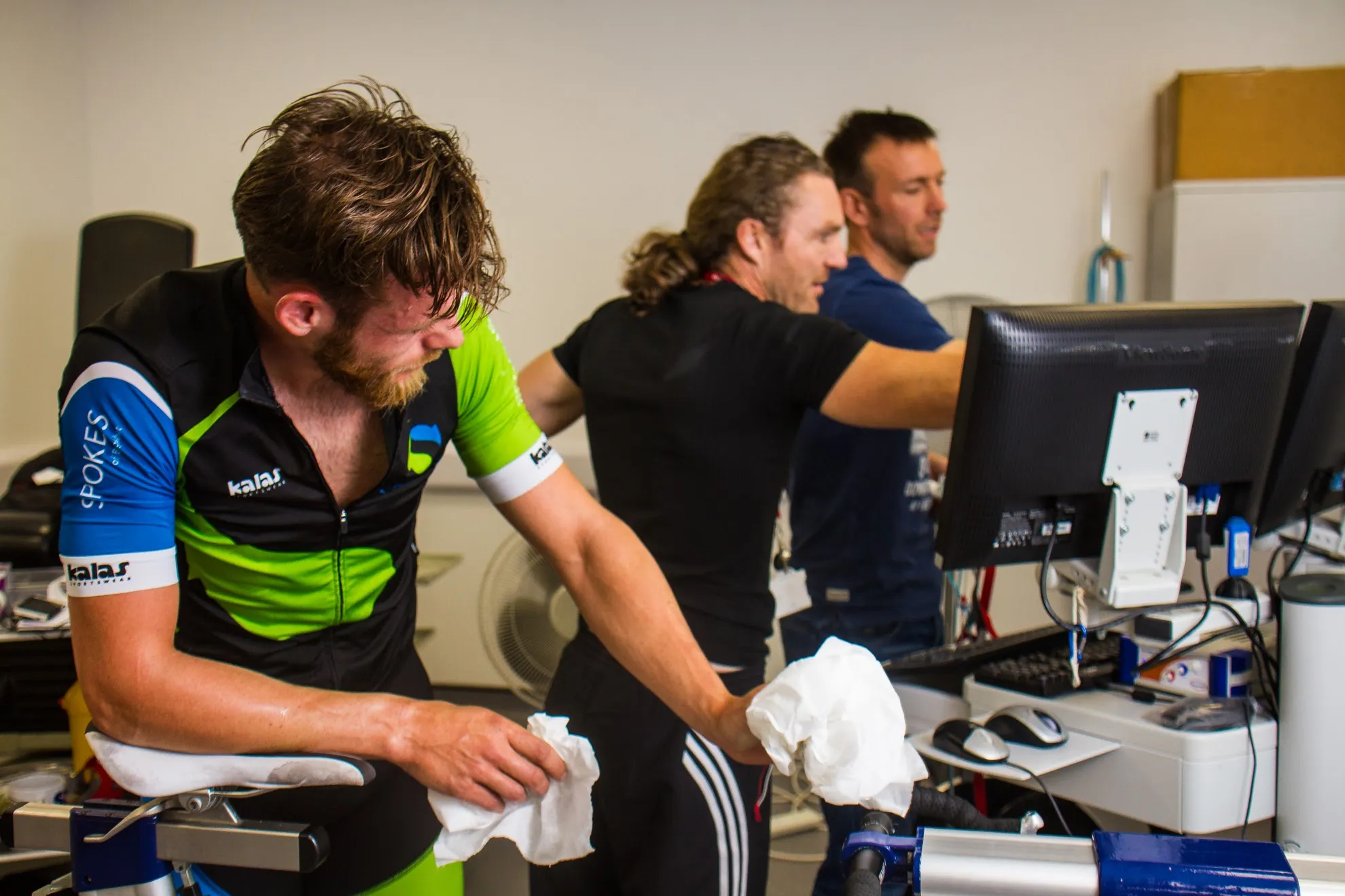 A cyclist on an indoor trainer being assisted by teammates