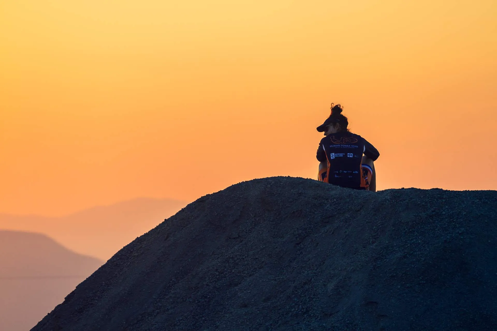 A lone silhouetted figure on a dark mound against an orange sunset sky