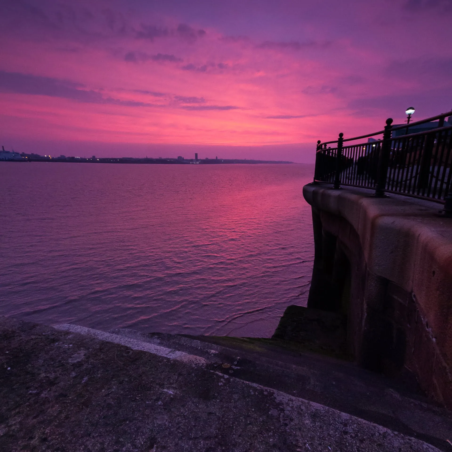 A vivid pink and purple sunset over the water from a promenade railing
