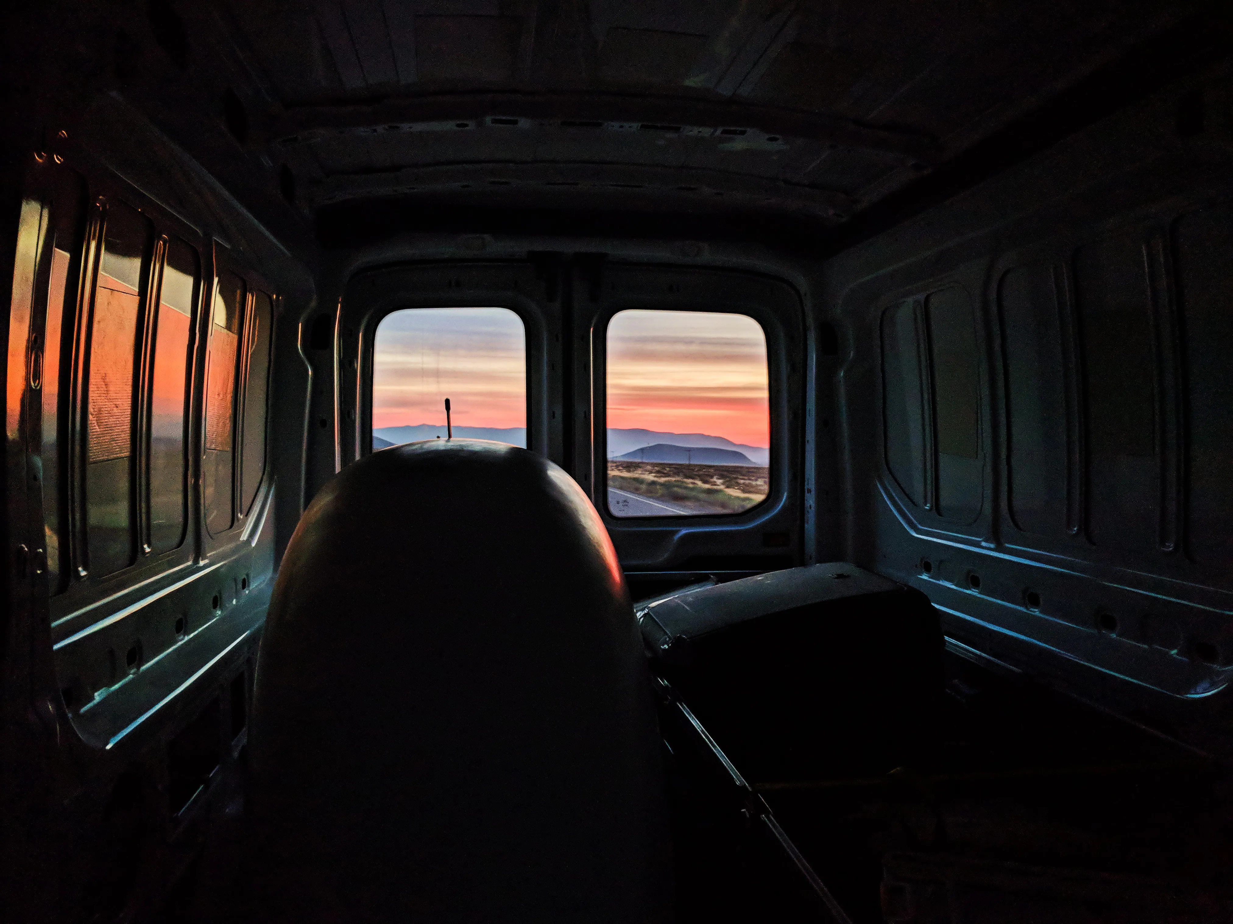 View through the rear windows of a van at a sunset-lit desert road and mountains